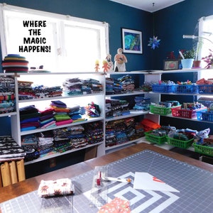 May include: A craft room with white shelves filled with colorful fabric scraps. The shelves are labeled "Where the magic happens!" There is a large cutting mat on a table in the foreground.