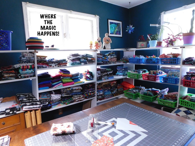 May include: A craft room with white shelves filled with colorful fabric scraps. The shelves are labeled "Where the magic happens!" There is a large cutting mat on a table in the foreground.