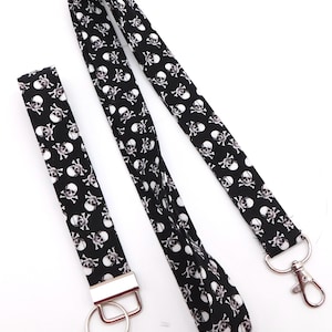 Könnte beinhalten: Schwarzes Schlüsselband und Armband-Set mit sich wiederholendem Muster aus weißen Totenköpfen. Das Schlüsselband hat einen Metall-Drehhaken, das Armband einen Metall-Schlüsselring. Ideal für Schlüssel oder einen Ausweis.