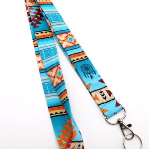 Op de afbeelding: Een blauwe en oranje gepatroneerde lanyard met een zilveren draaibare sluiting en een zilveren sleutelhanger. Het patroon bevat geometrische vormen, veren en een droomvanger.