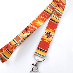 Op de afbeelding: Een kleurrijk koord met een geometrisch patroon in tinten oranje, geel, blauw en bruin. Het koord heeft een zilveren draaibare clip en een zilveren sleutelring.