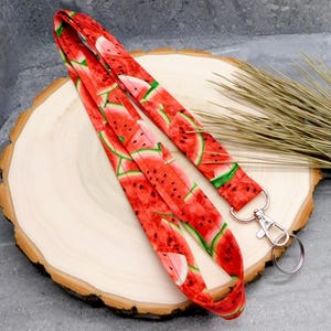 May include: A red lanyard with a repeating pattern of watermelon slices. The lanyard has a silver-colored metal clasp and key ring. The fabric is a bright red with green and black accents. The lanyard is laying on a wooden surface.