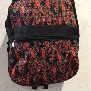 May include: Black backpack with a repeating pattern of red and black figures. The backpack has a black zippered front pocket and black shoulder straps. The main body of the backpack is covered in a repeating pattern of red and black figures.