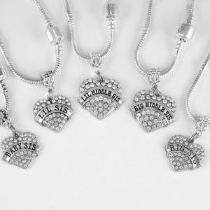 May include: Set of five silver necklaces with heart-shaped charms. Each charm has a different inscription: "Baby Sis", "Lil Sis", "Lil Middle Sis", "Big Middle Sis", and "Big Sis".