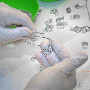 Puede incluir: Una pulsera de alambre extensible en tono plateado con un dije en forma de coraz&oacute;n incrustado con pedrer&iacute;a transparente y la palabra CHEER grabada en el centro. La pulsera se sostiene con manos enguantadas en blanco y hay m&aacute;s dijes visibles.