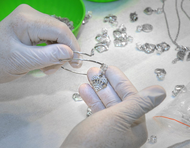 Puede incluir: Una pulsera de alambre extensible en tono plateado con un dije en forma de coraz&oacute;n incrustado con pedrer&iacute;a transparente y la palabra CHEER grabada en el centro. La pulsera se sostiene con manos enguantadas en blanco y hay m&aacute;s dijes visibles.