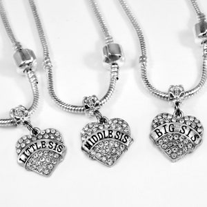 Puede incluir: Tres collares de cadena de serpiente plateada, cada uno con un colgante en forma de coraz&oacute;n incrustado con pedrer&iacute;a transparente. Una banda sobre cada coraz&oacute;n muestra el texto LITTLE SIS, MIDDLE SIS o BIG SIS en letras negras.
