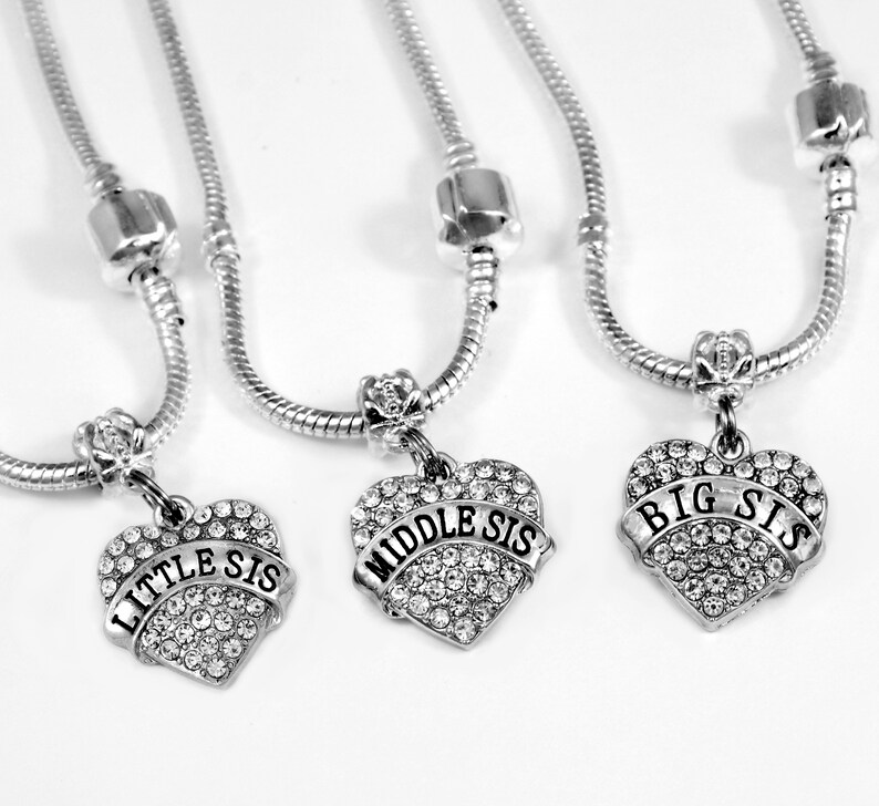 Puede incluir: Tres collares de cadena de serpiente plateada, cada uno con un colgante en forma de coraz&oacute;n incrustado con pedrer&iacute;a transparente. Una banda sobre cada coraz&oacute;n muestra el texto LITTLE SIS, MIDDLE SIS o BIG SIS en letras negras.