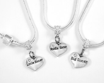 May include: Three silver necklaces with heart-shaped charms. Each charm has a rhinestone and the words "Big Sister", "Middle Sister", and "Little Sister" engraved on them.