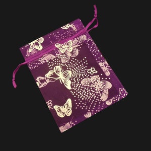 Puede incluir: Una bolsa rectangular de color morado con cierre de cord&oacute;n, decorada con un patr&oacute;n repetitivo de mariposas blancas y peque&ntilde;os puntos blancos. La bolsa de tela tiene un cord&oacute;n morado a juego en la parte superior, anudado en el lado izquierdo.