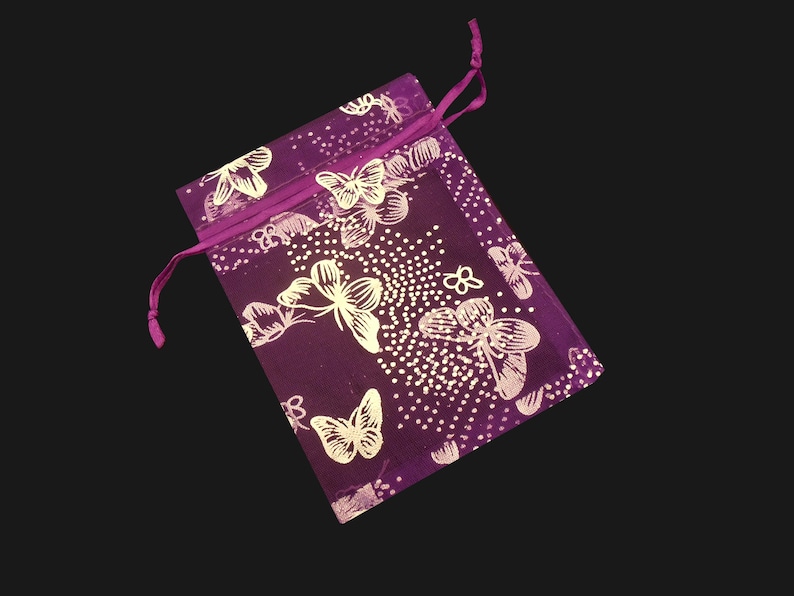 Puede incluir: Una bolsa rectangular de color morado con cierre de cord&oacute;n, decorada con un patr&oacute;n repetitivo de mariposas blancas y peque&ntilde;os puntos blancos. La bolsa de tela tiene un cord&oacute;n morado a juego en la parte superior, anudado en el lado izquierdo.