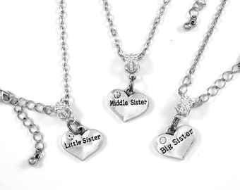 May include: Three silver necklaces with heart-shaped charms that say "Big Sister", "Middle Sister", and "Little Sister". Each charm has a small rhinestone.