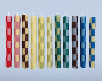 May include: Eleven cylindrical taper candles arranged in a row, each featuring a distinct two-tone chequered pattern. The candles display various colour combinations including red, pink, brown, yellow, green, and blue, all topped with a white wick.