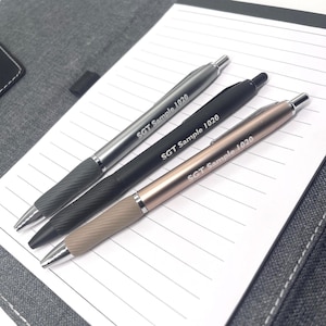 May include: Three ballpoint pens in silver, black, and rose gold, each with a textured grip and the text "SGT Sample 1020." The pens are lying on a lined notepad, with a gray fabric background.