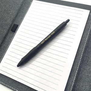 May include: A black pen with the text "SGT Sample 1020" printed on the side rests on a white lined notepad. The notepad is inside a gray fabric cover with a black elastic band. The pen is angled across the notepad.