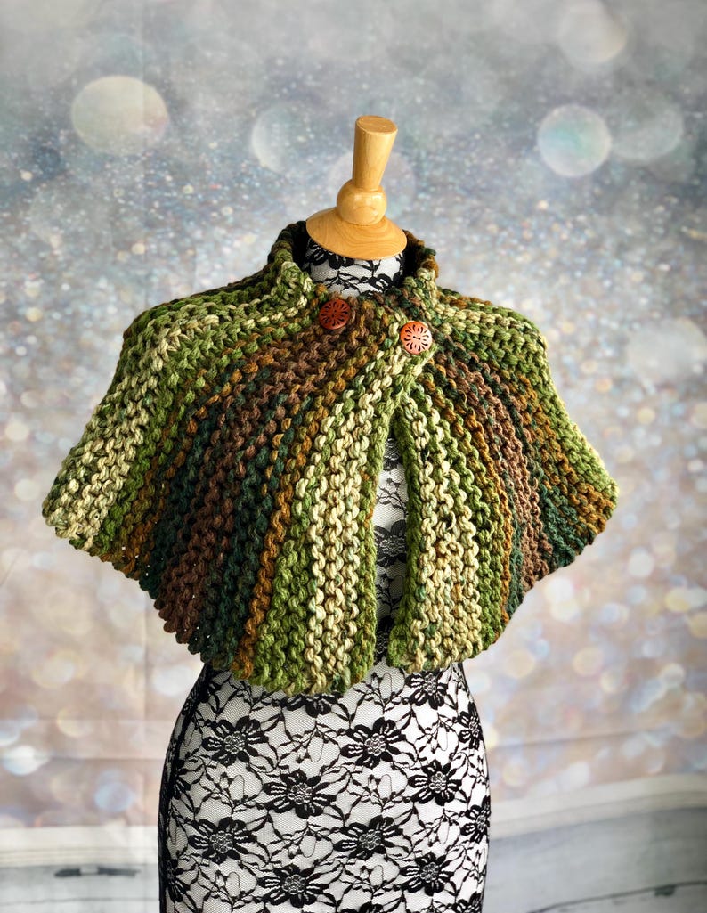 May include: A green, brown, and beige knitted cape with wooden buttons. The cape is on a white dress form.