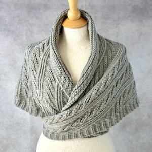 May include: A grey knitted shawl with a cable knit pattern. The shawl is draped over a mannequin.