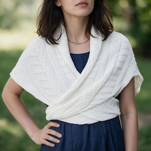 May include: A white knitted wrap featuring a textured cable pattern and a wide shawl collar. The garment is designed to cross over the chest, creating a layered appearance with a soft, woven texture throughout the fabric of the wrap.