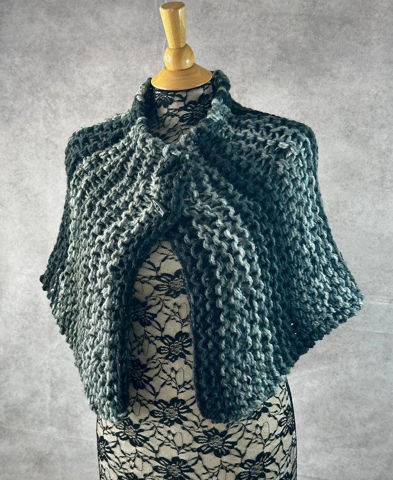 May include: A dark green and gray knitted shawl with a textured pattern. The shawl is draped over a mannequin.
