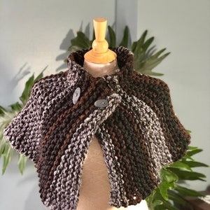 May include: A brown, gray, and white knitted shawl with two buttons. The shawl is on a wooden mannequin.