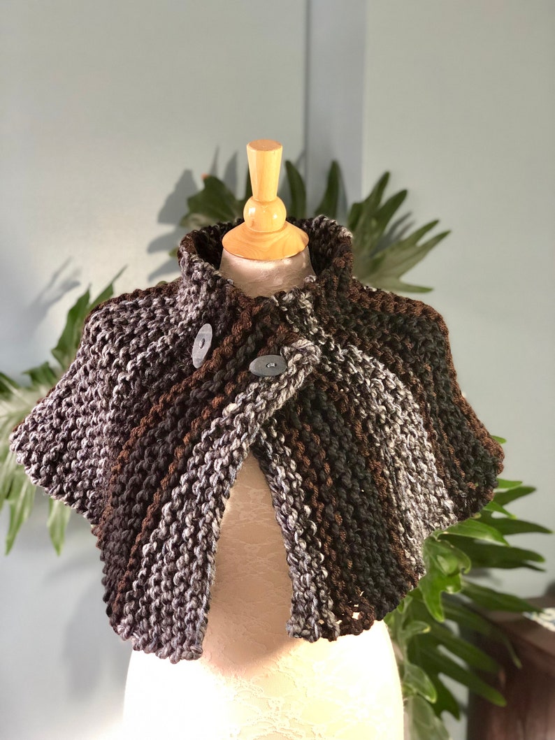 May include: A brown, gray, and white knitted shawl with two buttons. The shawl is on a wooden mannequin.