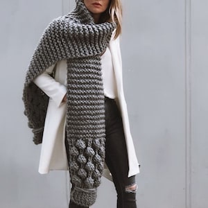 May include: A gray knitted scarf with a textured pattern and large, round, gray pom-poms. The scarf is draped around a person's neck and shoulders.