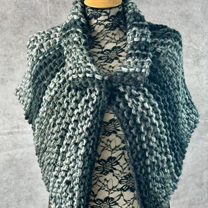 May include: A green and gray knitted shawl with a textured pattern. The shawl is draped over a mannequin.