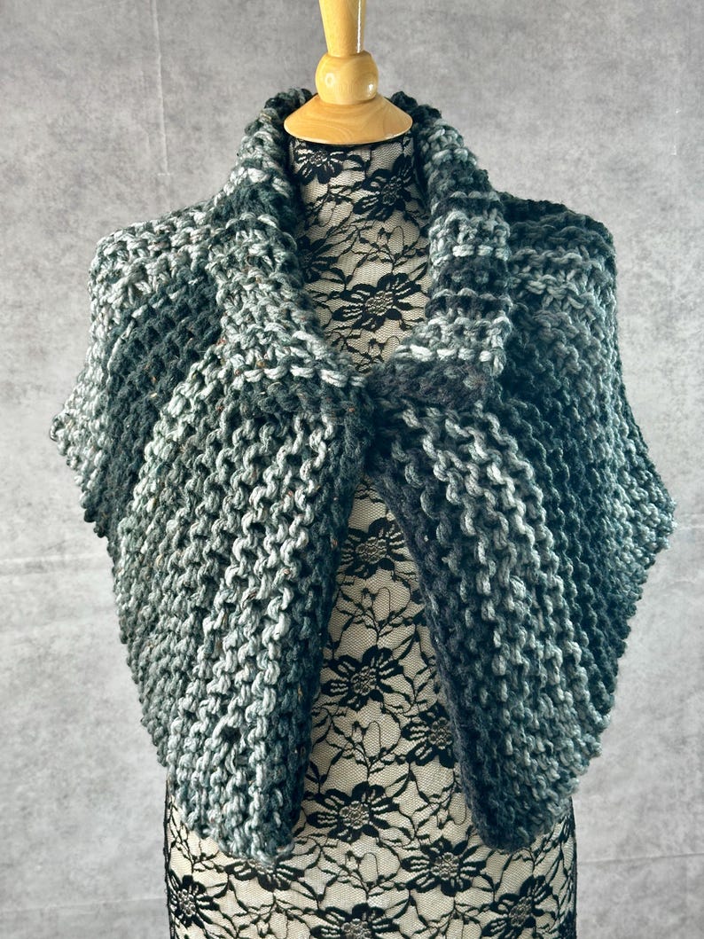 May include: A green and gray knitted shawl with a textured pattern. The shawl is draped over a mannequin.
