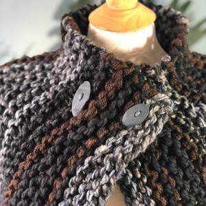 May include: A brown, black, and gray knitted cape with a high collar and two large buttons. The cape is made of a chunky yarn and has a textured look.