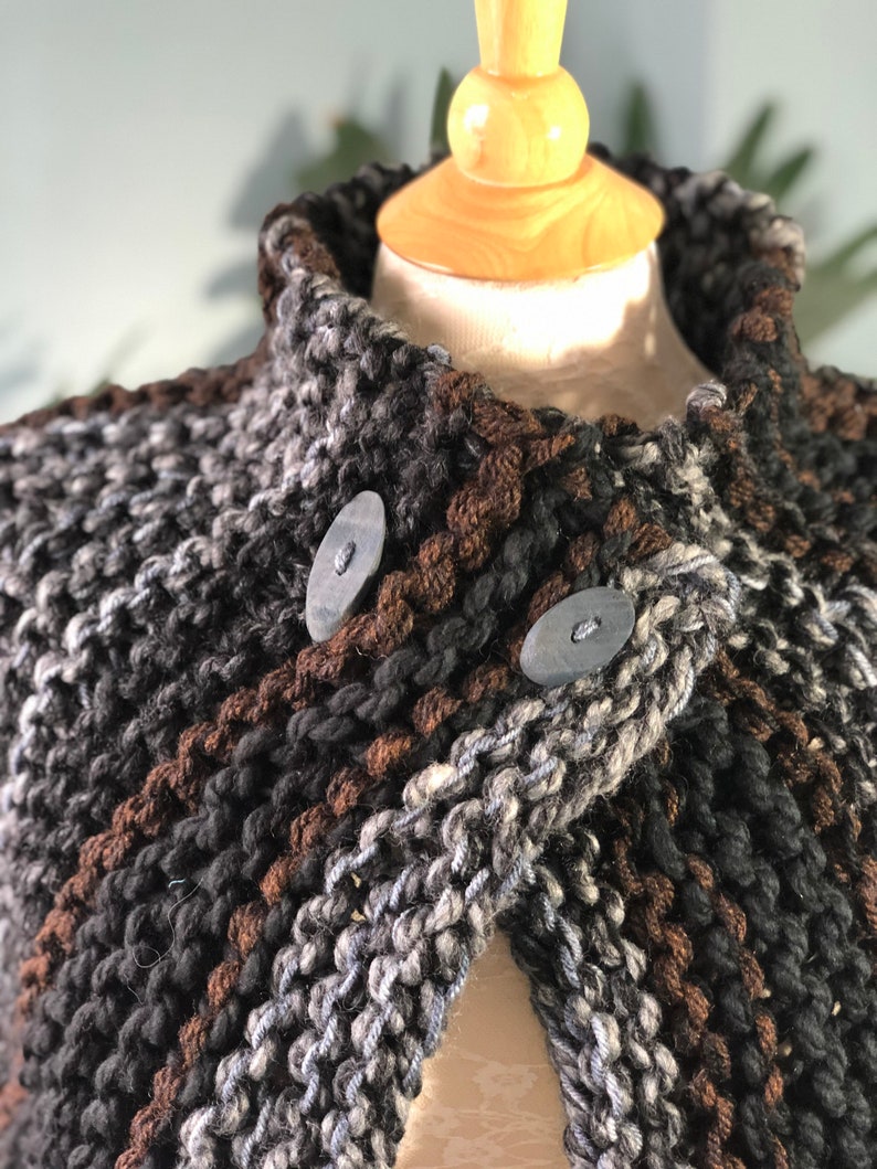 May include: A brown, black, and gray knitted cape with a high collar and two large buttons. The cape is made of a chunky yarn and has a textured look.