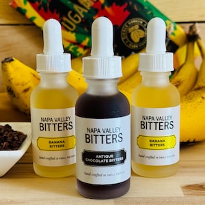 May include: Three glass bottles of Napa Valley Bitters, with labels for Banana Bitters, Antique Chocolate Bitters, and Banana Bitters. The bottles are on a wooden surface with bananas and cocoa nibs in the background.