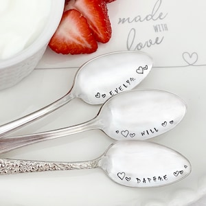 May include: Three silver spoons with personalized names, "Evelyn", "Will", and "Daphne", engraved on the front of each spoon. The spoons are arranged on a white surface with strawberries and a bowl of yogurt in the background. The text "made with love" is printed on the surface.