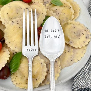 May include: A fork and spoon with engraved text, next to a plate of ravioli. The fork reads "DREW" with a heart symbol. The spoon reads "MY ONE & RAVI-ONLY". The ravioli is covered in a creamy sauce, with tomatoes and basil.