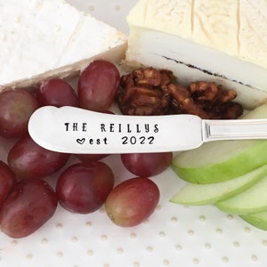May include: A silver cheese knife with the text "THE REILLYS est 2022" engraved on the handle. The knife is lying on a white plate with grapes, cheese, walnuts, and apple slices.