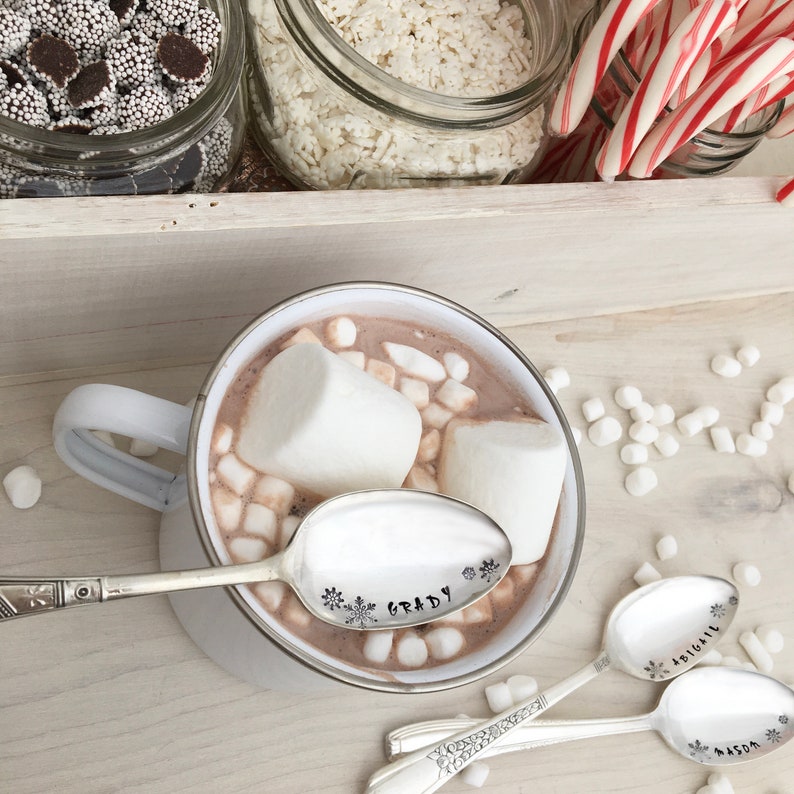 May include: A white mug filled with hot chocolate and marshmallows, with a silver spoon engraved with the name "Grady" and snowflake designs.  Three additional spoons are visible in the background, engraved with the names "Abigail", "Mason", and "Grady".