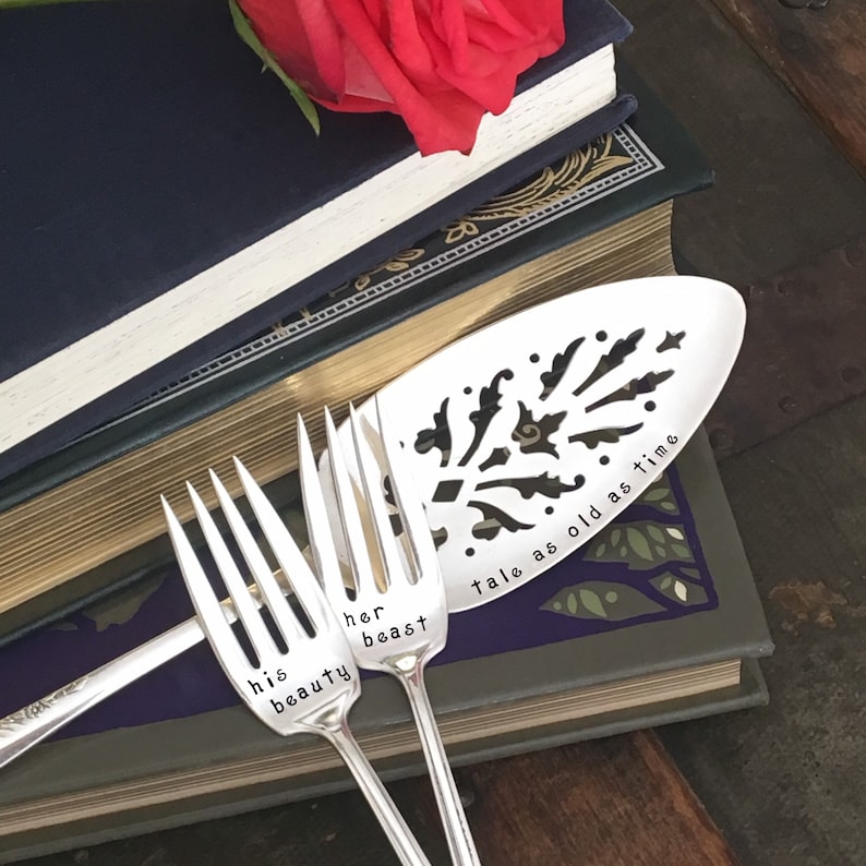 May include: Three silver utensils with engraved text. The top utensil is a serving spoon with the text "tale as old as time". The two forks have the text "his beauty" and "her beast".