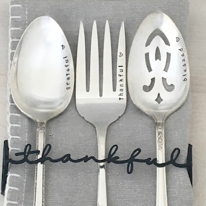 Puede incluir: Tres utensilios de plata con las palabras "grateful", "thankful" y "blessed" grabadas en ellos. Los utensilios están unidos por un soporte de metal negro con la palabra "thankful" escrita en él.