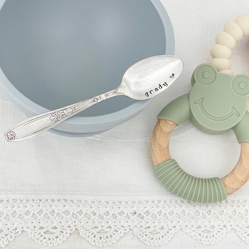 May include: A silver baby spoon with the name "Grady" engraved on it, next to a light blue bowl and a green and wood baby teether.