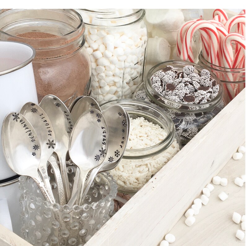 May include: A white wooden tray with various toppings for hot chocolate. The toppings include mini marshmallows, white chocolate chips, candy sprinkles, and candy canes. There are also six silver spoons with engraved names.