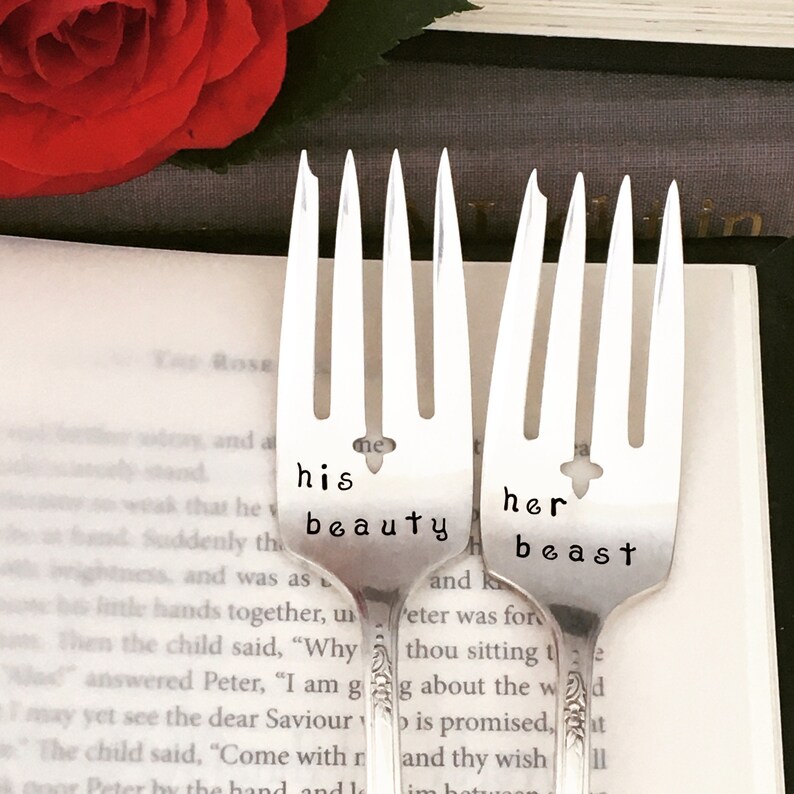 May include: Two silver forks with the words "his beauty" and "her beast" engraved on them. The forks are placed on a book with a red rose in the background.