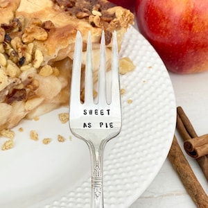 May include: A silver fork with the words "Sweet as Pie" engraved on the handle. The fork is on a white plate with a slice of apple pie and a red apple.