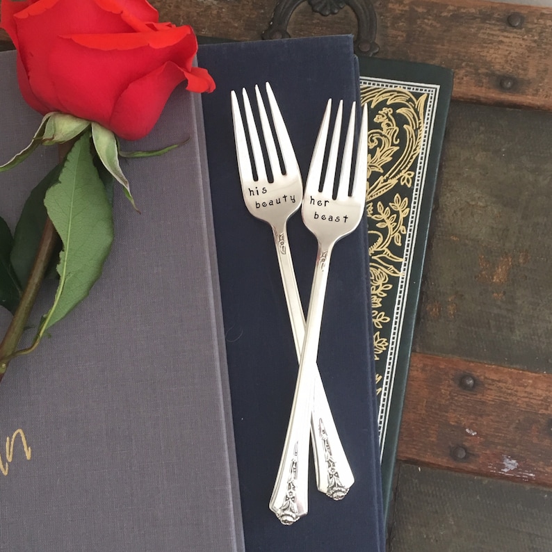 May include: Two silver forks with the words "his beauty" and "her beast" engraved on them. The forks are crossed and resting on a stack of books.