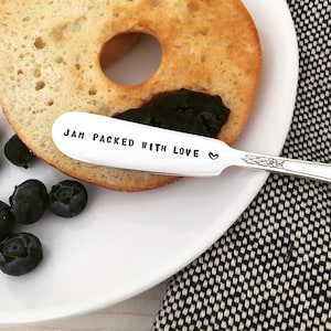 May include: A silver butter knife with the words "JAM PACKED WITH LOVE" engraved on the handle. The knife is resting on a white plate with a bagel and blueberry jam.