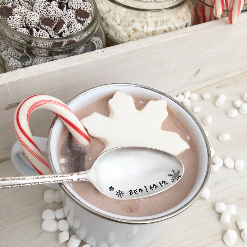 May include: A white ceramic mug filled with hot chocolate topped with a large white marshmallow shaped like a snowflake. A silver spoon with the engraved text "Benjamin" and a snowflake rests on the rim of the mug.