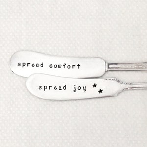 May include: Two silver butter knives with engraved sayings. The top knife reads "spread comfort". The bottom knife reads "spread joy" with two stars.