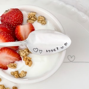 Pu&ograve; includere: Un cucchiaio d'argento con il nome "Abbey" inciso su di esso, circondato da cuori. Il cucchiaio &egrave; in una ciotola di yogurt con fragole e granola.