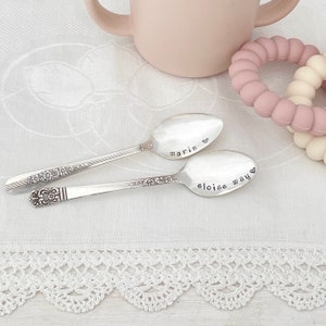 May include: Two silver spoons with engraved names, "Marin" and "Eloise May", on a white crocheted tablecloth. The spoons are a thoughtful gift for a new baby.