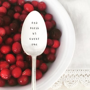 May include: A silver spoon with the words "God Bless Us Every One" engraved on it. The spoon is resting in a bowl of red cranberries.