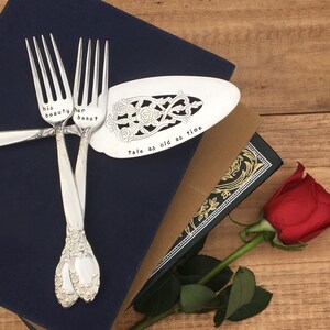 May include: A set of three silver utensils with engraved text, including a cake server with the words "tale as old as time" and two forks with the words "his beauty" and "her beast". The utensils are on top of a blue book with a rose in the foreground.