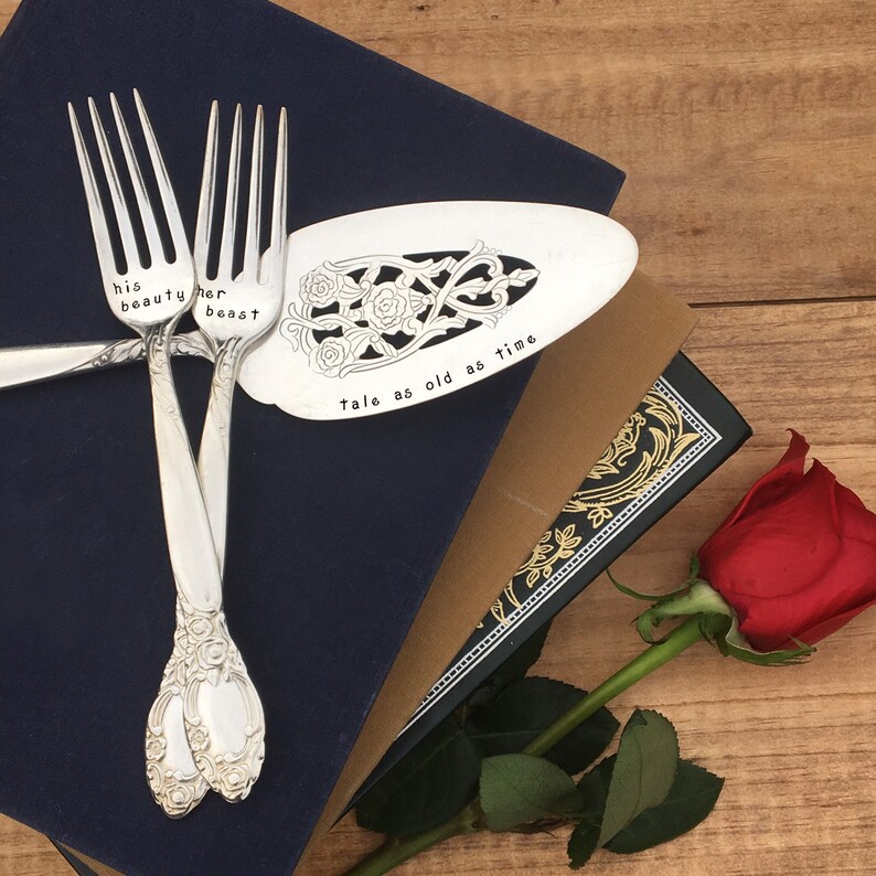 May include: A set of three silver utensils with engraved text, including a cake server with the words "tale as old as time" and two forks with the words "his beauty" and "her beast". The utensils are on top of a blue book with a rose in the foreground.